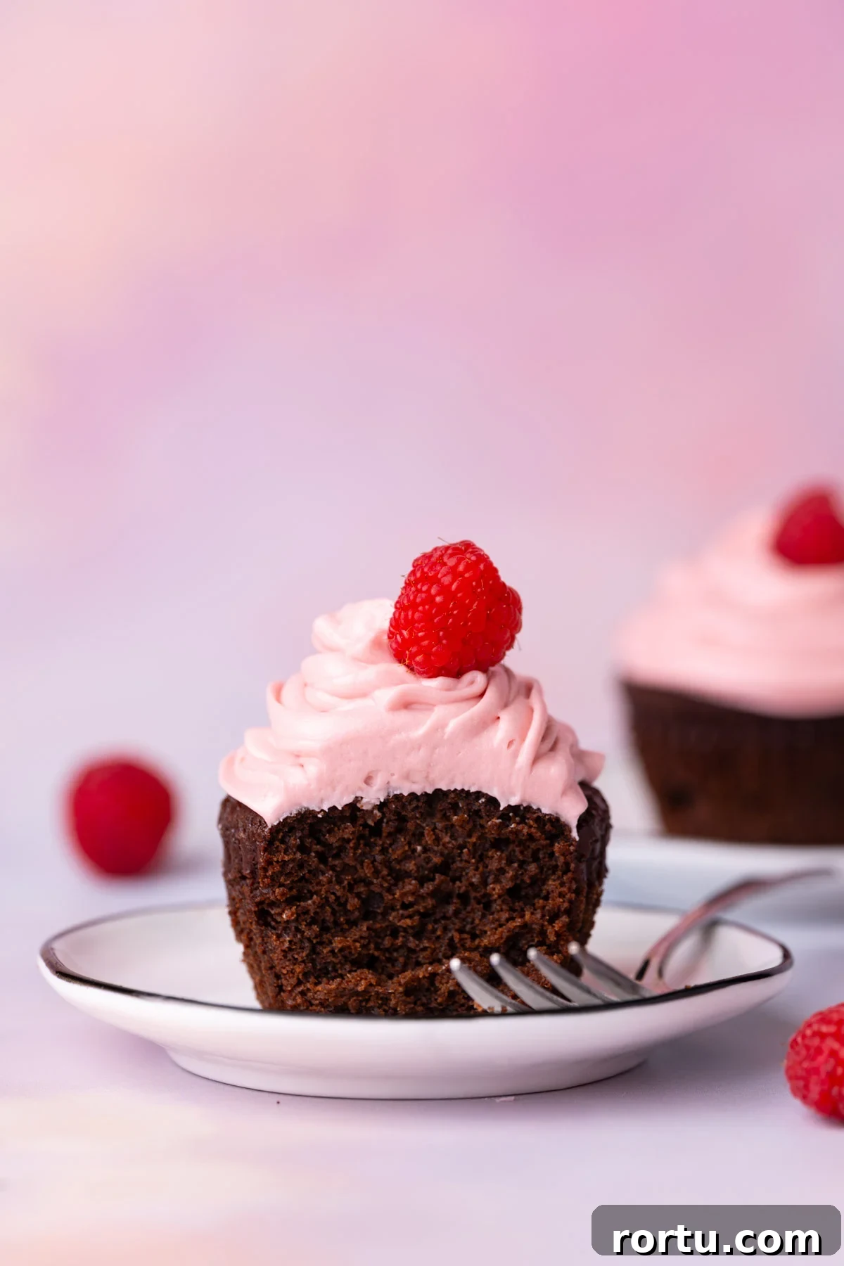 Chocolate cupcake with raspberry buttercream topped with a fresh raspberry with forkful taken out.