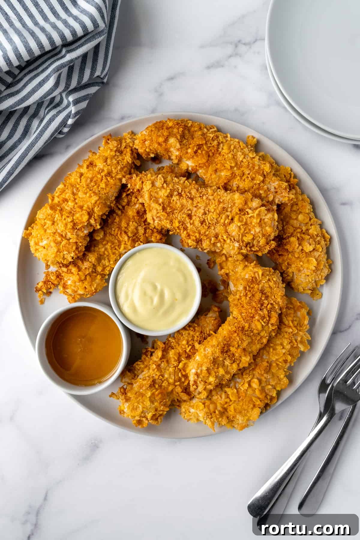Crispy Baked Cornflake Chicken Tenders Recipe 6 Baked Cornflake Chicken Tenders on a plate with dipping sauces.