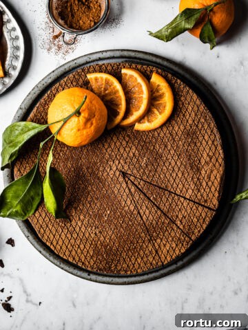 A chocolate orange cheesecake decorated with orange slices and a whole orange with a few slices removed, on a white marble surface. There are more oranges with leaves nearby, a small bowl of chopped chocolate, and a plate of sliced candied oranges.