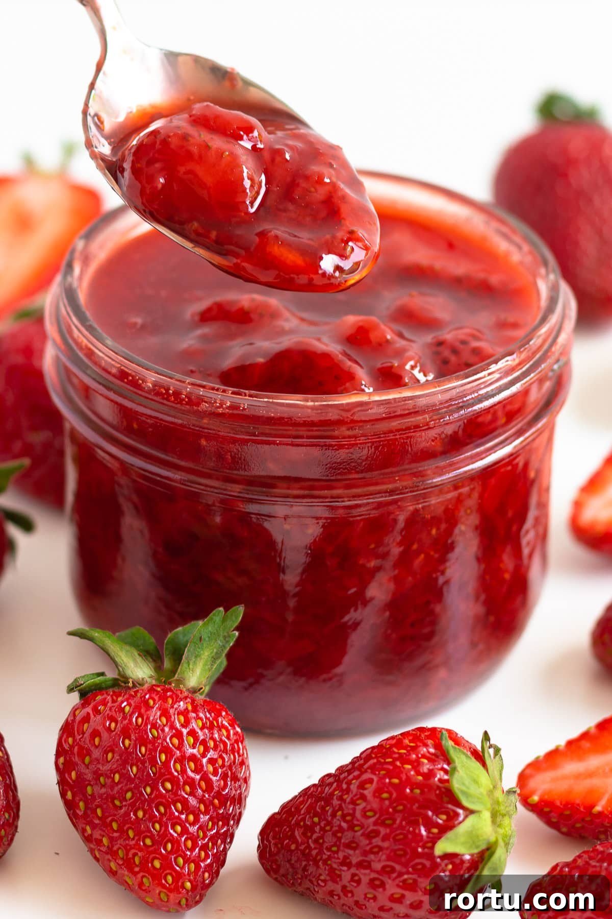 Jar of strawberry compote surrounded by fresh strawberries and spoon scooping some out.