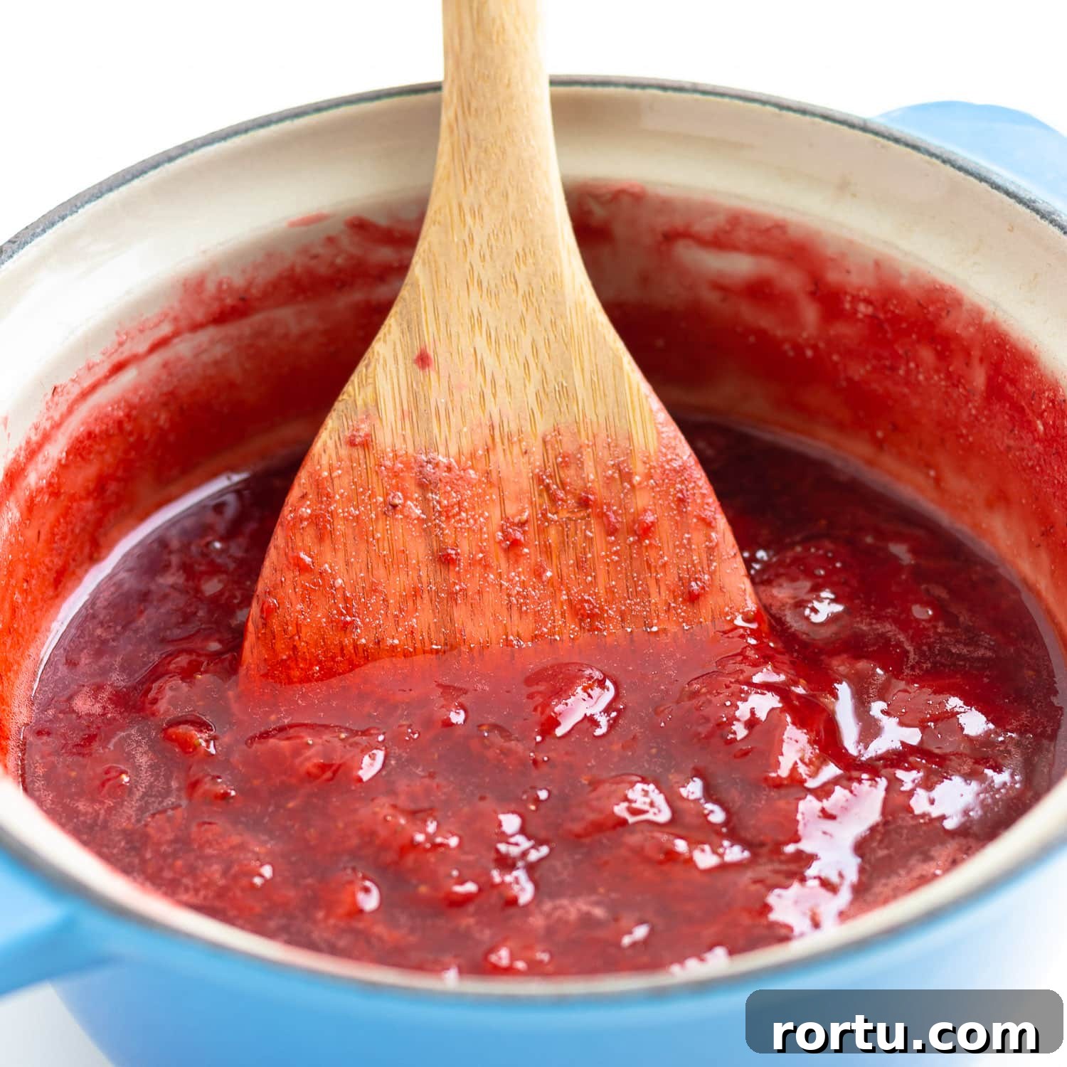 Wooden spoon stirring strawberry compote in saucepan.