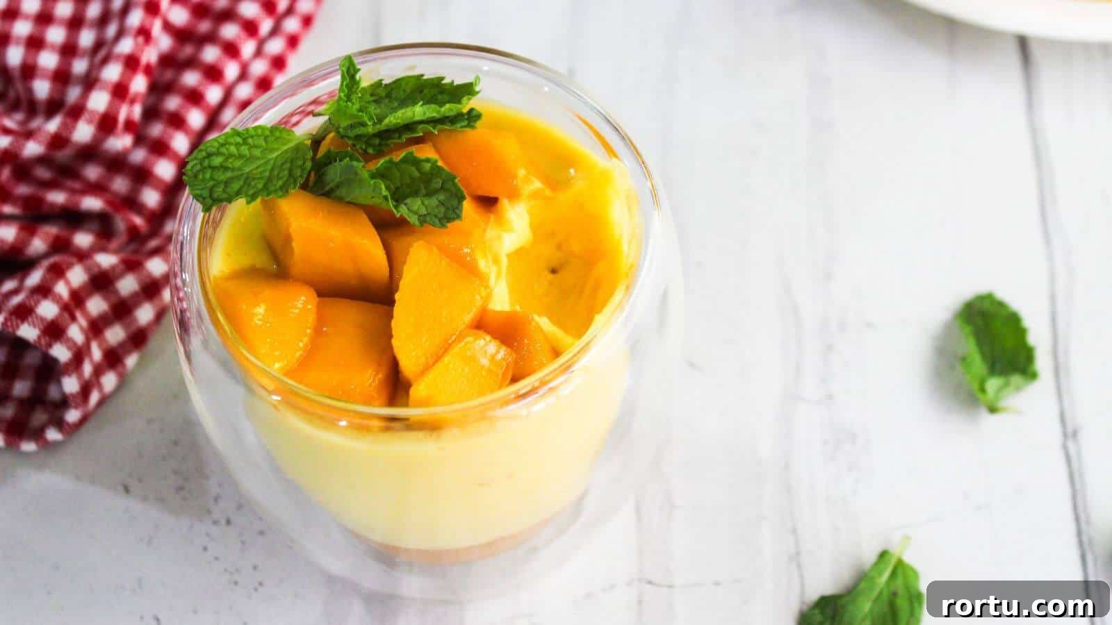 A glass cup of mango mousse topped with fresh mango cubes and mint leaves sits on a white table next to a red checkered cloth and a plate of sliced mango.