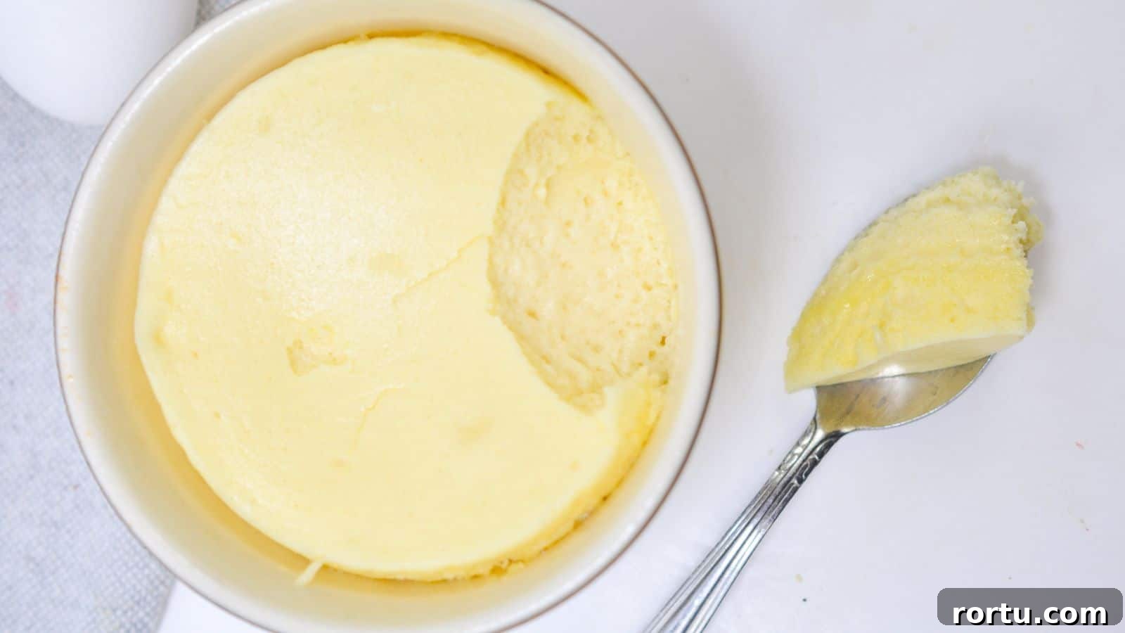 A ramekin of creamy, smooth cheesecake with a spoon resting beside it, taking out a scoop on a white background.