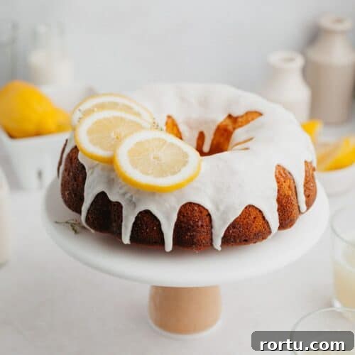 A lemon bundt cake covered in a white glaze and slices of lemon on a white cake stand.