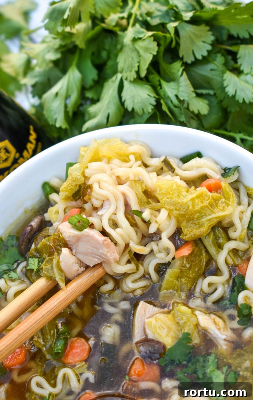 wooden chop sticks lifting noodles out of a white bowl filled with Asian chicken noodle soup with cabbage