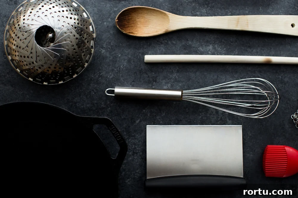kitchen equipment including whisk steamer wooden spoon bench scraper and cast iron skillet on dark background