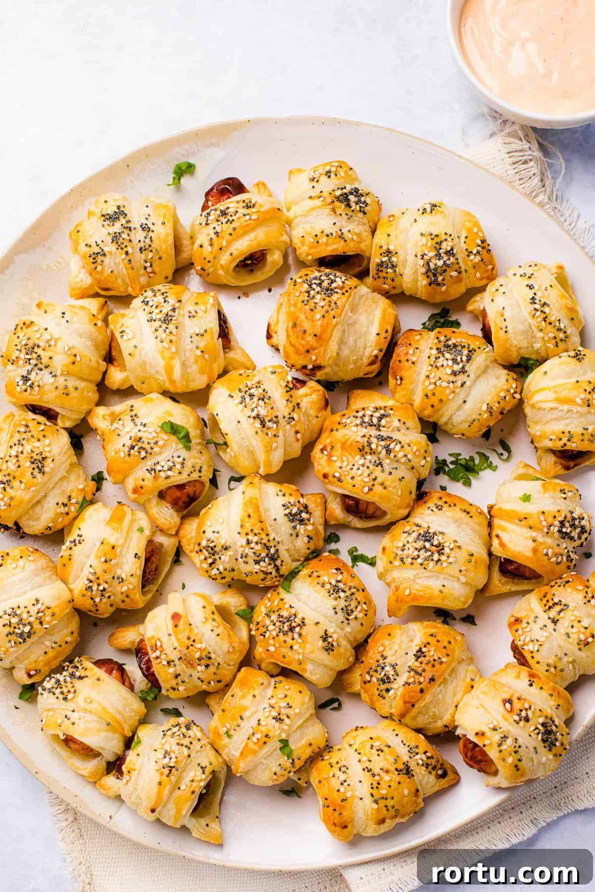 A serving platter holding about two dozen pigs in a blanket. A bowl of dipping sauce sits nearby.