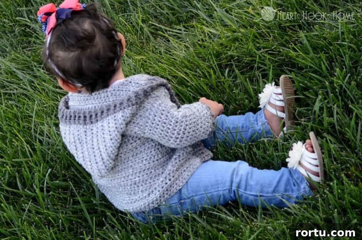 Image shows a baby sitting in the grass wearing a hoodie crochet grey sweater. -Skip To My Lou