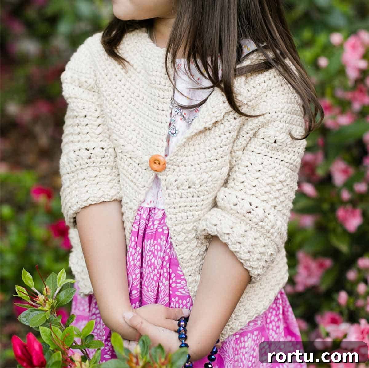Image shows a little girl wearing crochet cardigan in front of a blurry background. Skip To My Lou